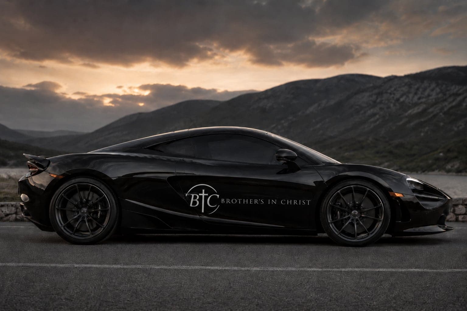 Black sports car with the Brothers In Christ logo on the door, parked on a mountain road at dusk