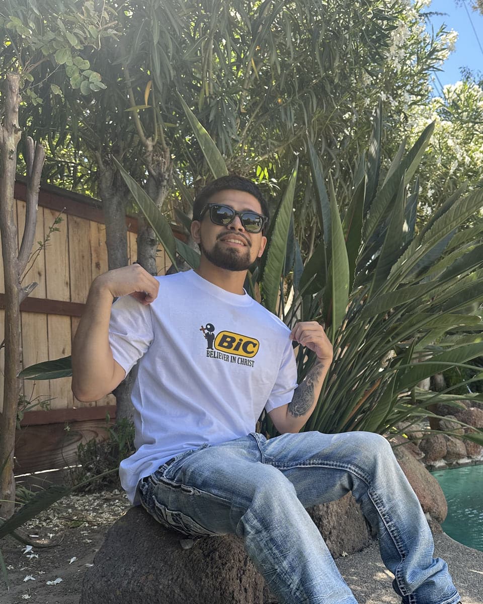 Marc Anthony Flores-Vega in a white Believer In Christ tee and sunglasses, seated on a rock in a garden
